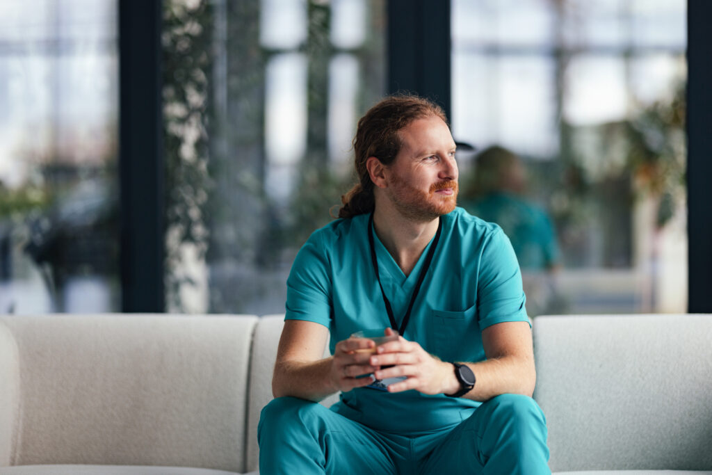 Male nurse sitting on a couch taking a break and looking thoughtfully out the window.